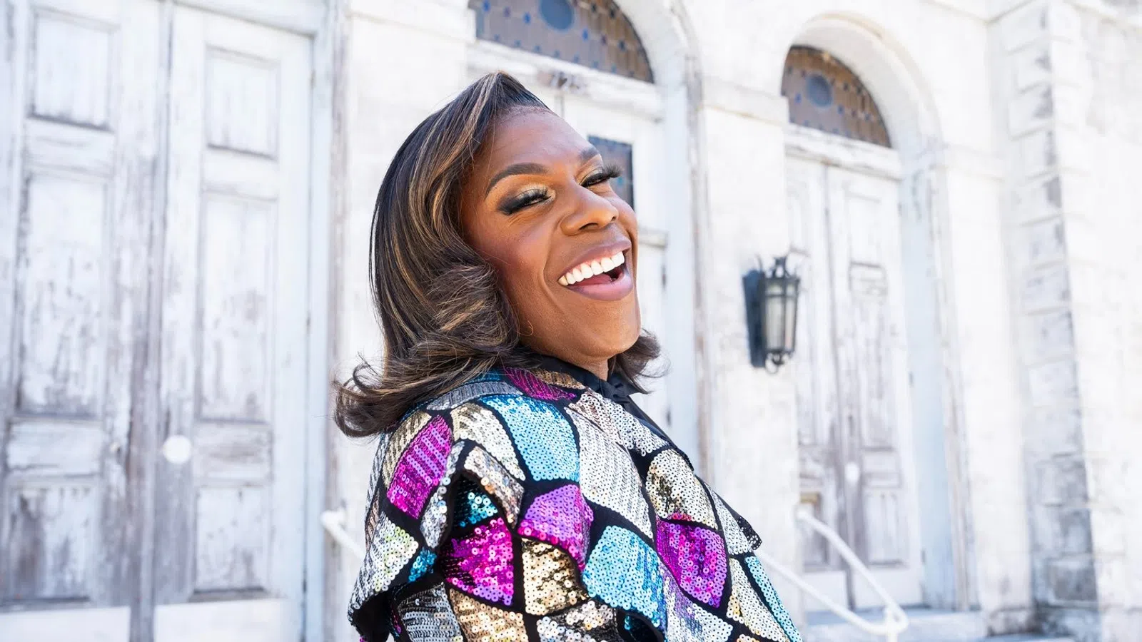 Smiling person in colorful sequin jacket posing outdoors near weathered white doors and stone steps