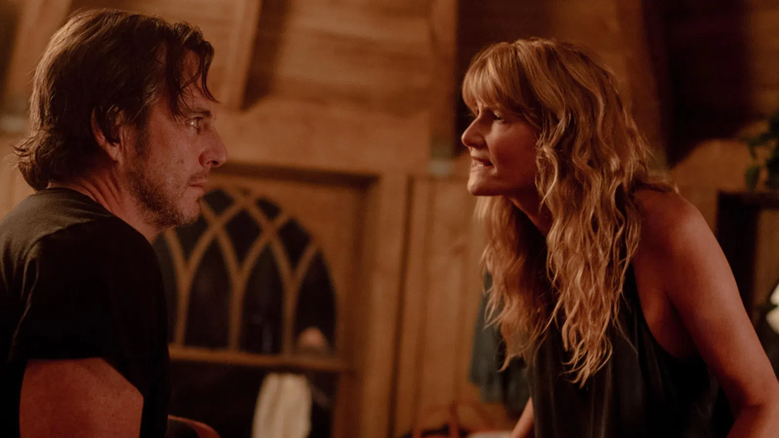 Man and woman engage in an intense conversation inside a warmly lit wooden cabin at night.