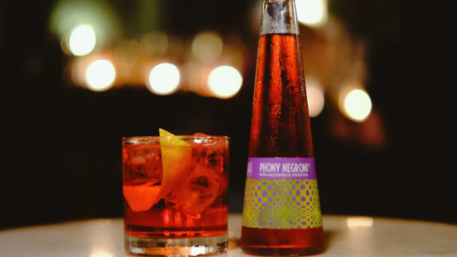 Bottle of Phony Negroni non-alcoholic cocktail next to a glass with ice and orange peel on a table.