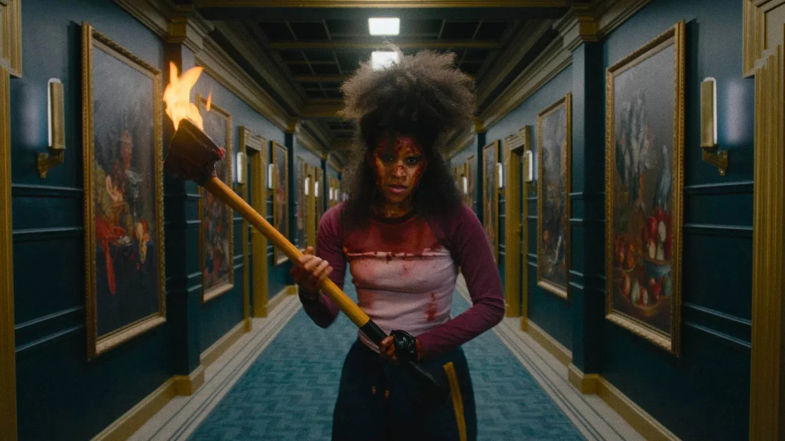 Person with blood-stained clothes holding a flaming torch standing in a dimly lit hallway lined with paintings.