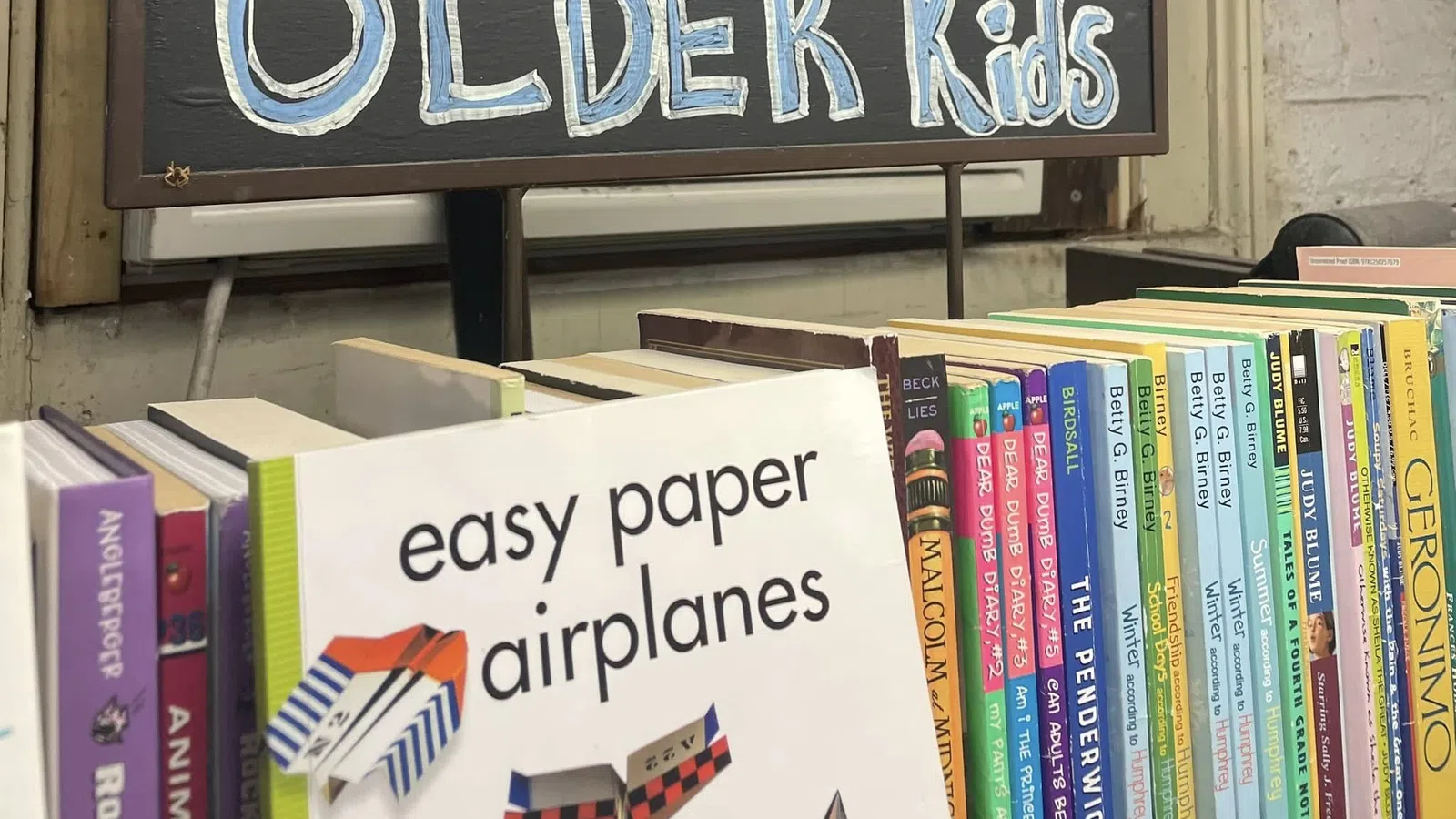 Books for older kids displayed on a shelf with a sign reading Older Kids and a book about easy paper airplanes.