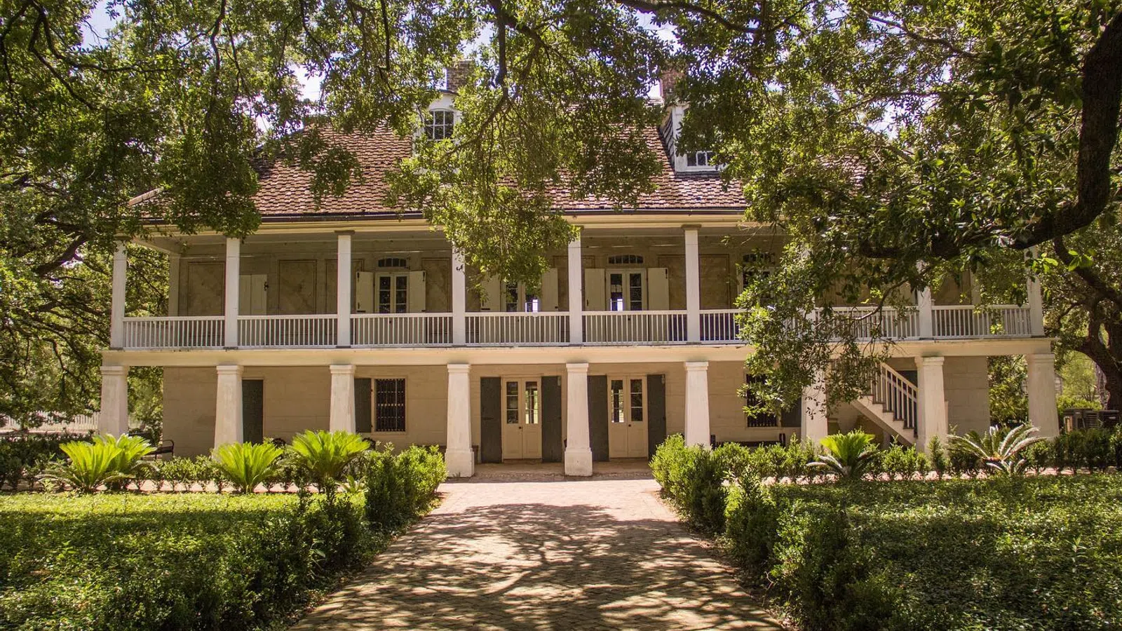 Historic two-story plantation house surrounded by lush greenery and shaded by large oak trees.
