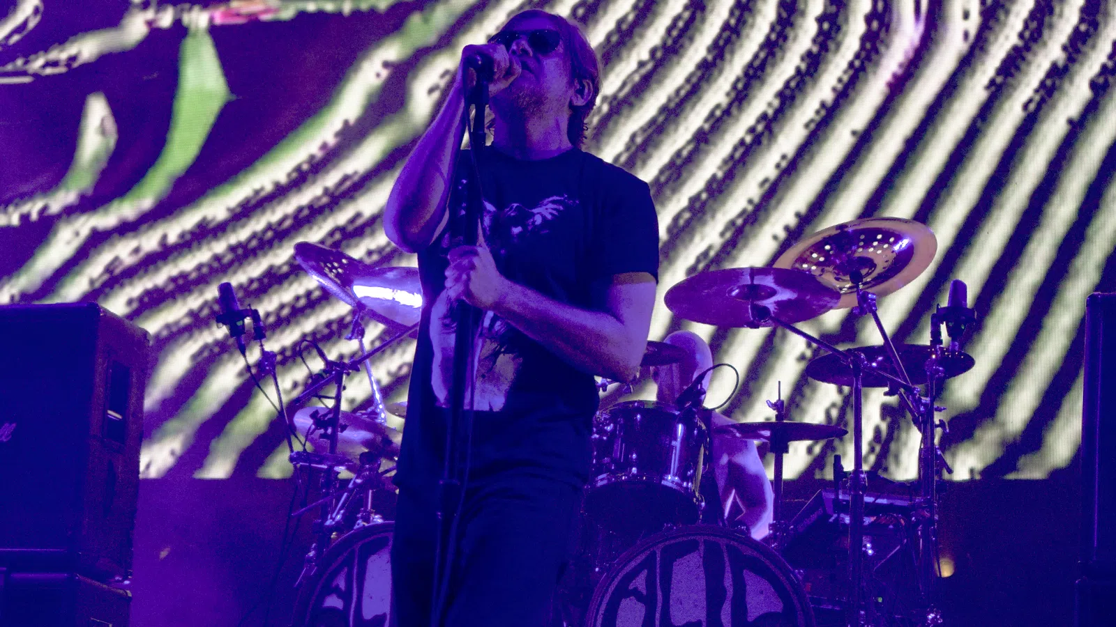 Male rock vocalist singing on stage with microphone and drummer performing behind during purple light concert.