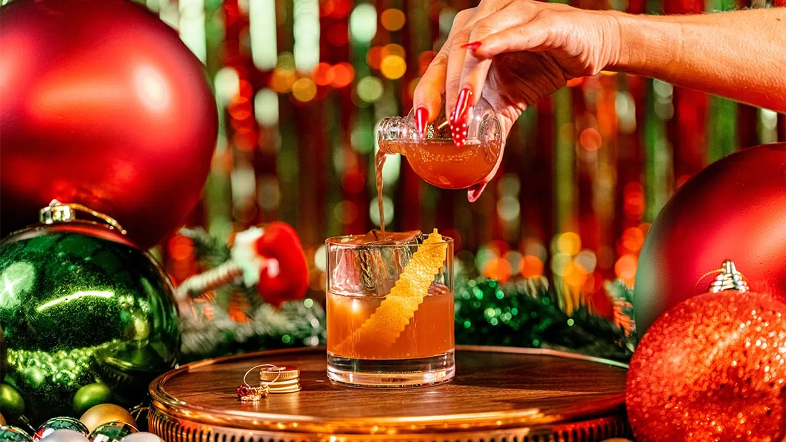 Hand pouring a festive cocktail with an orange twist on a wooden table surrounded by Christmas ornaments and lights.