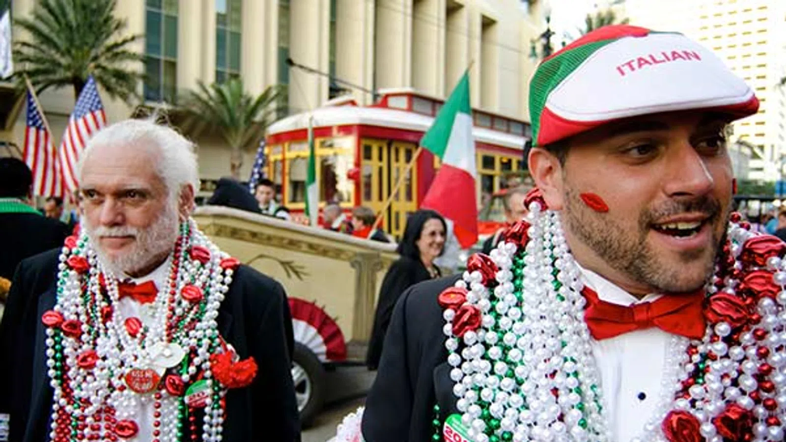 Two men wearing festive Italian-themed beads and accessories during a street celebration with flags and trolley.