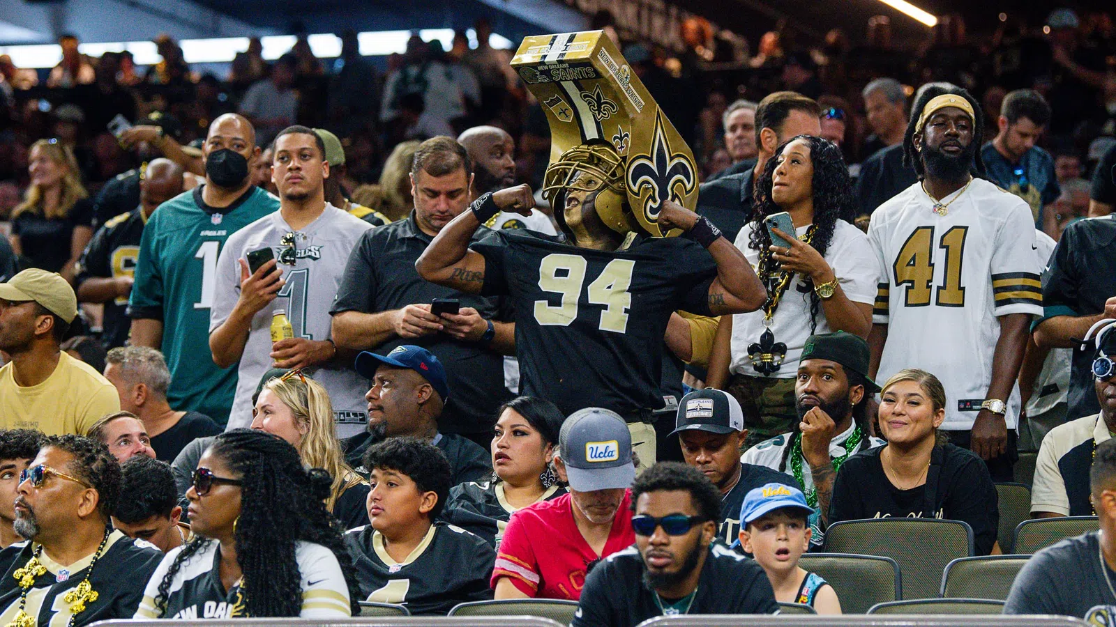 Fans wearing New Orleans Saints jerseys and gear cheer in a crowded stadium during a football game.