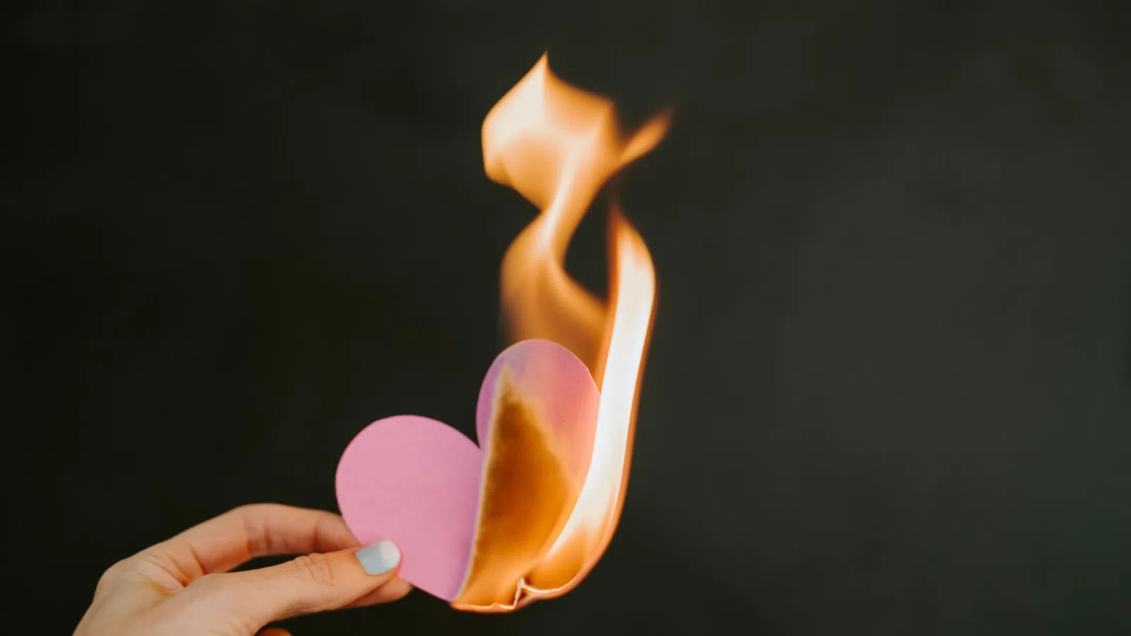 Hand holding a pink paper heart burning with bright orange flames against a dark background.