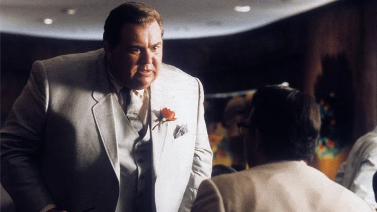 Man in a light suit with a rose boutonniere talks intensely to seated men in a dimly lit restaurant setting.