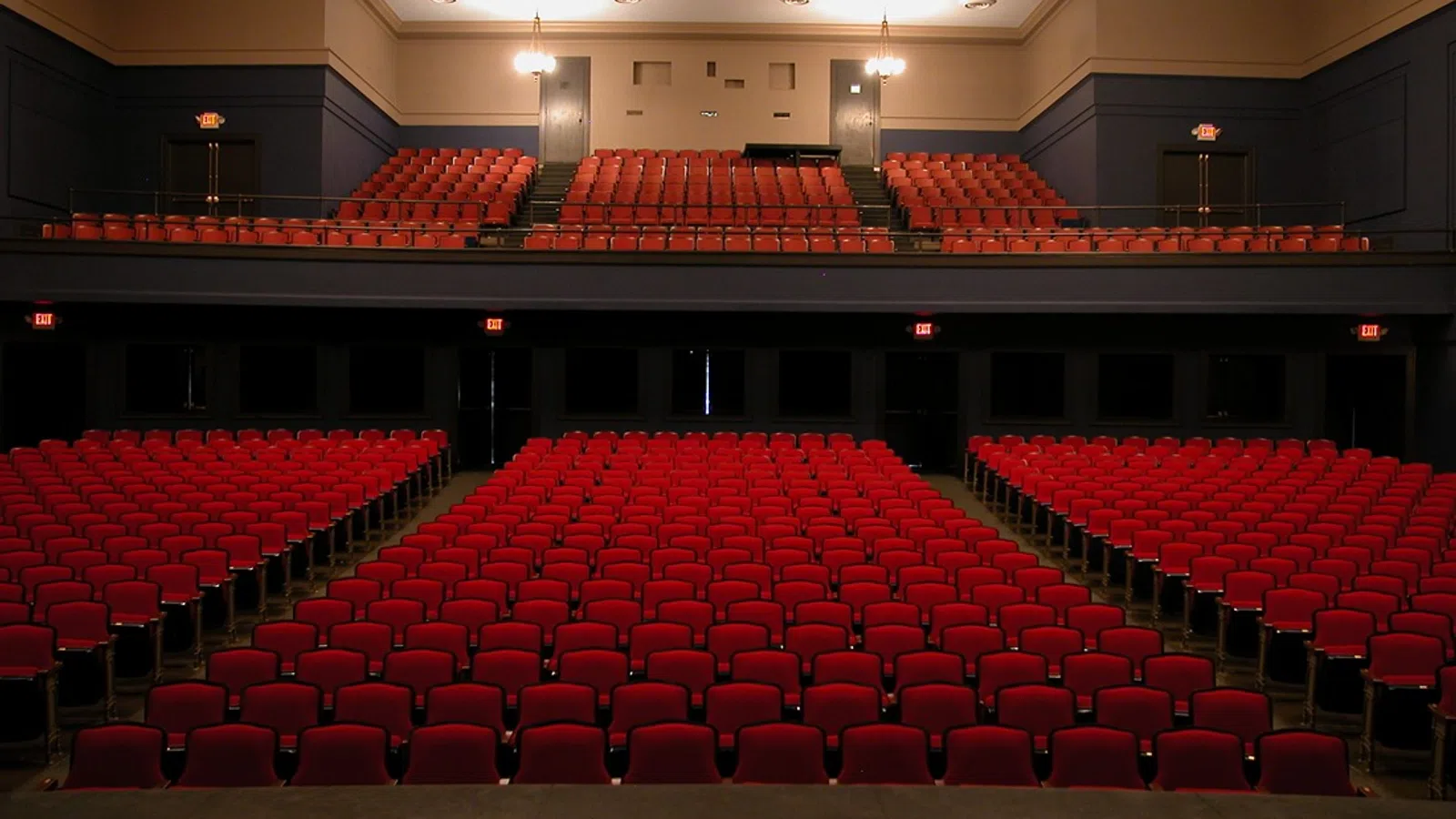 Empty theater with rows of red seats and a balcony, viewed from the stage, under warm lighting.