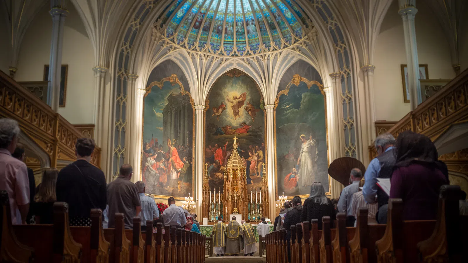 Congregation attending mass in a decorated Catholic church with stained glass dome and religious paintings