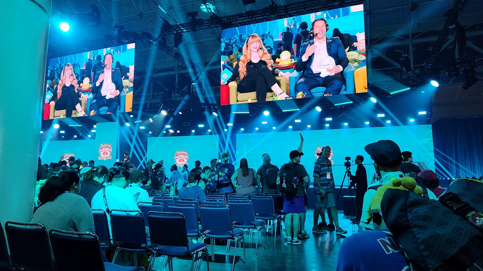 Crowded gaming event with audience watching live commentators on large screens under blue stage lighting.