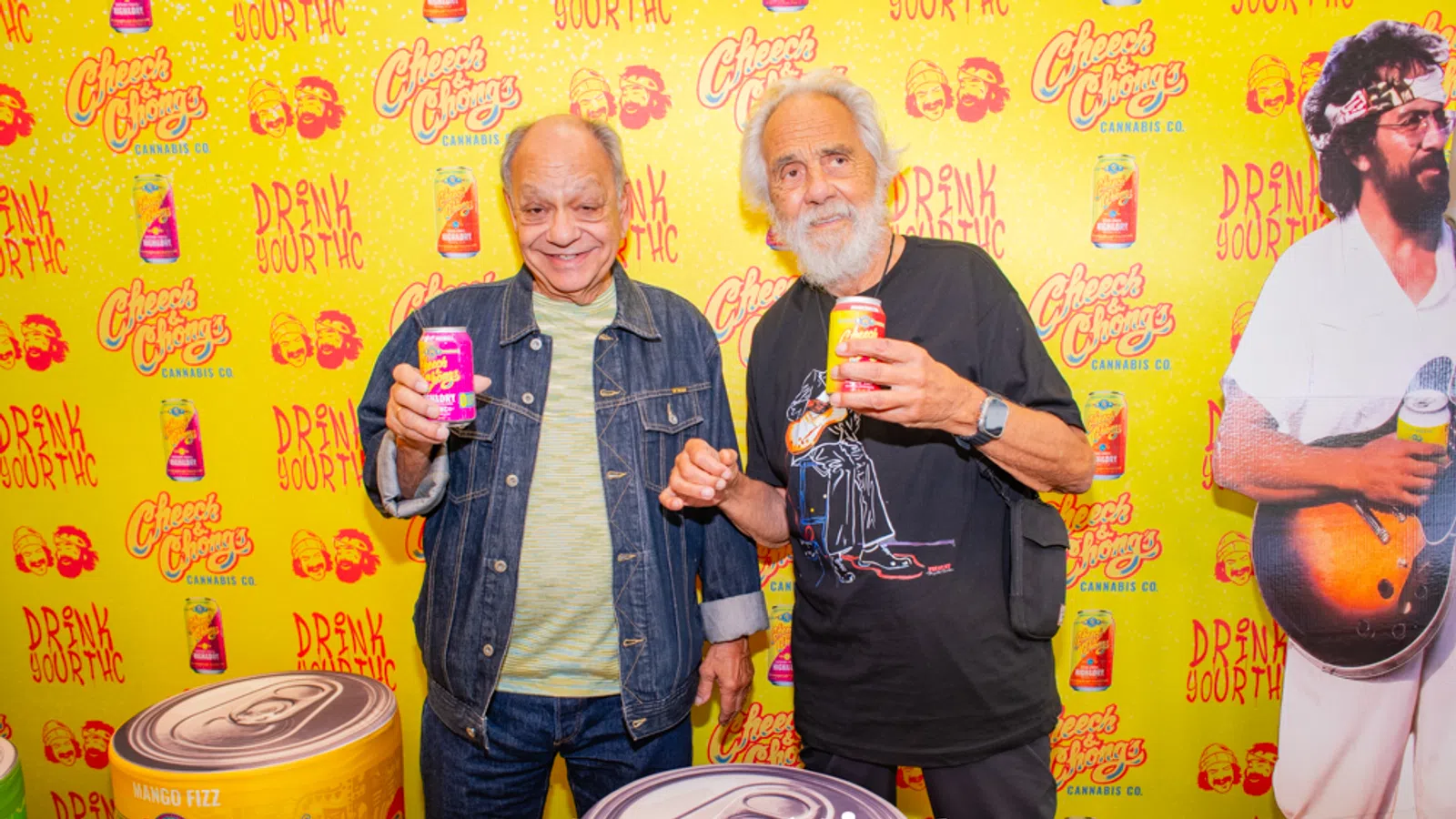 Two men smiling and holding cans of Cheech & Chong's cannabis drink in front of a branded yellow backdrop.