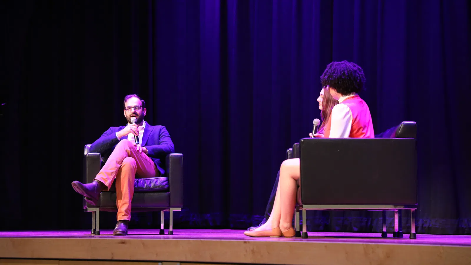 Man and two women seated on stage in armchairs having a discussion with microphones under purple stage lighting.