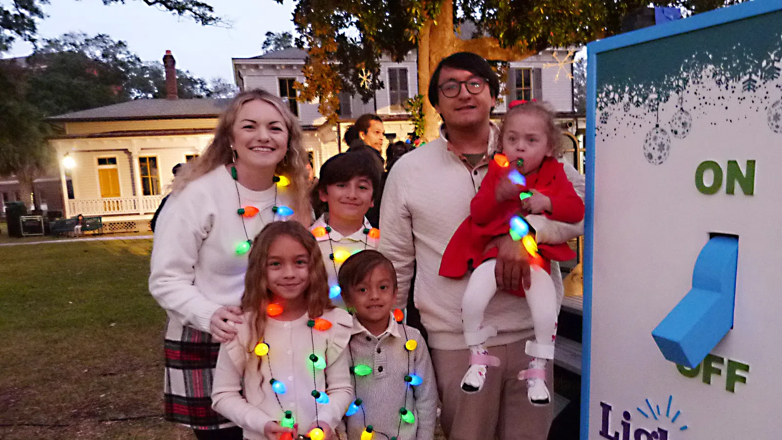 Family dressed in holiday lights standing outdoors near a giant on-off switch decoration at dusk
