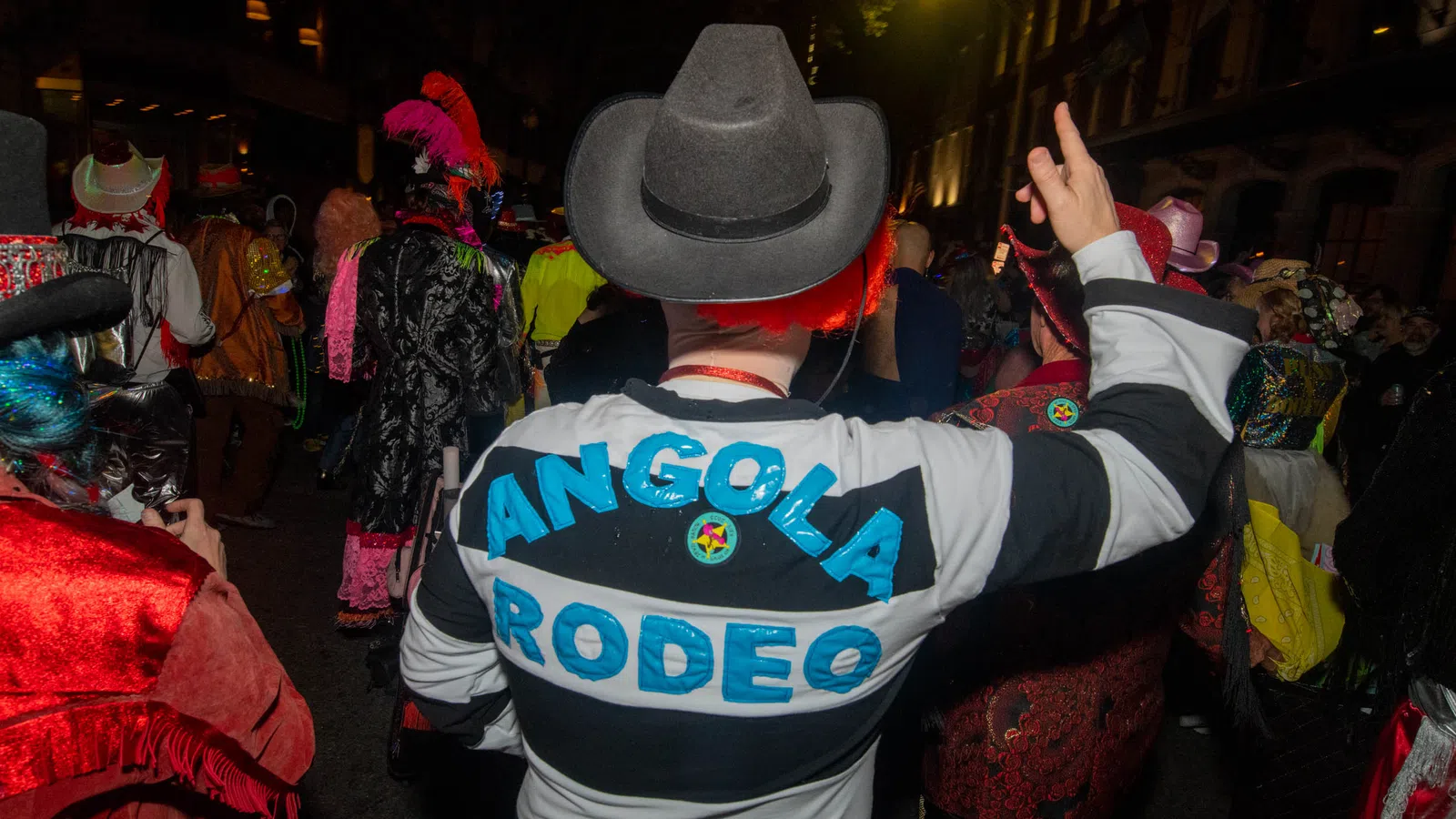 Person wearing Angola Rodeo striped shirt and cowboy hat at a lively nighttime costume event with colorful attendees.