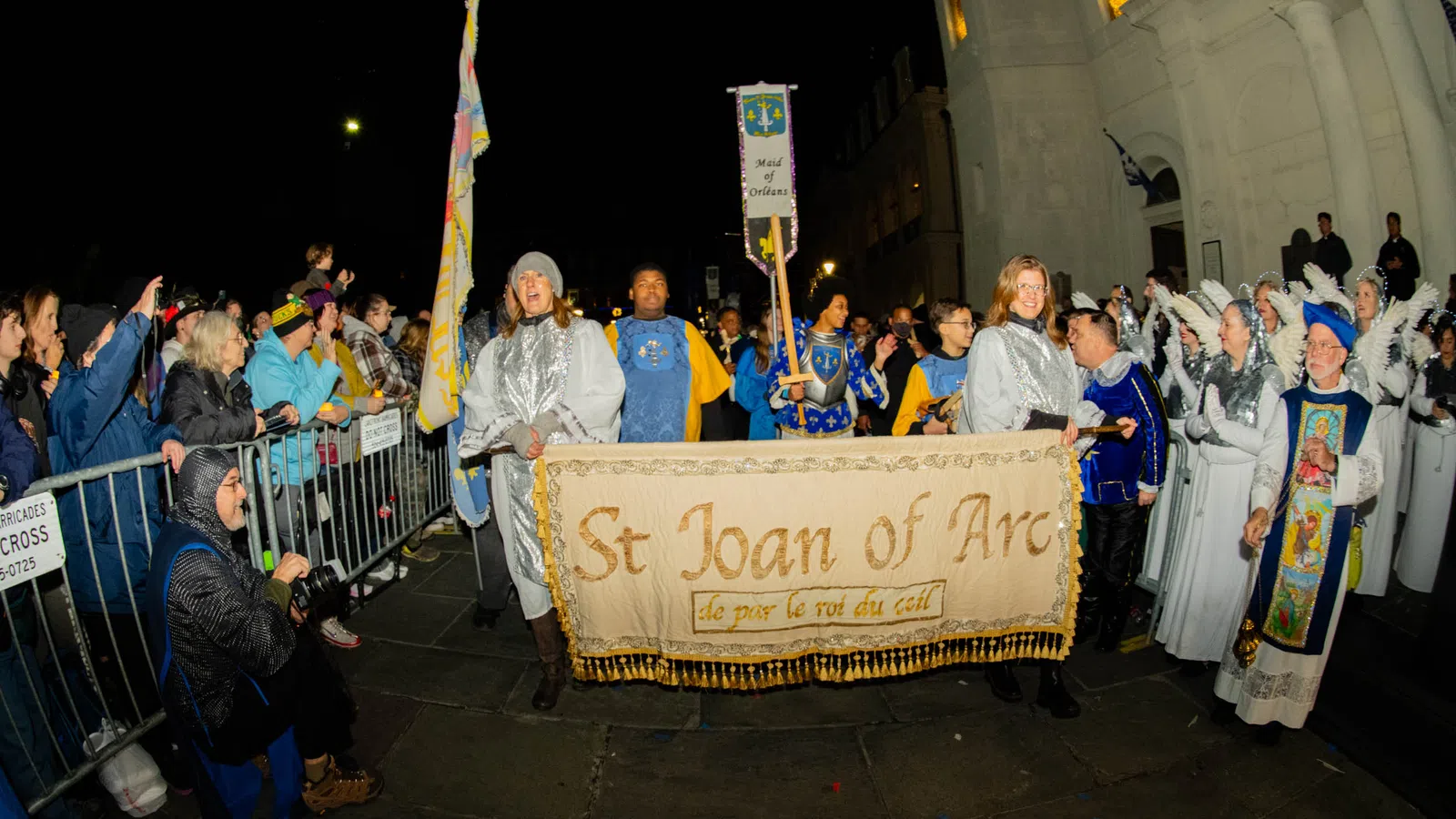 Night parade with people dressed as St Joan of Arc and medieval figures holding a banner and flags in front of a crowd.