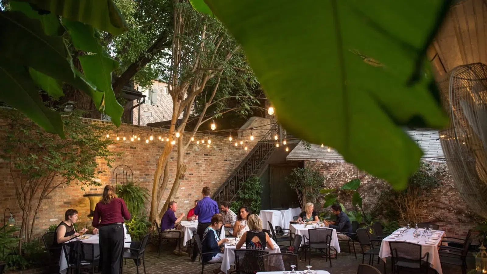Outdoor restaurant patio with diners, string lights, brick walls, and tropical leaves creating a cozy ambiance.