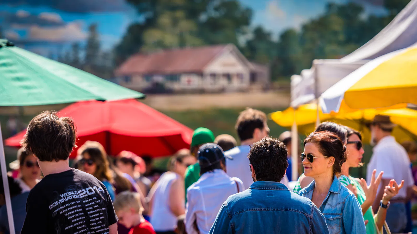 People socializing outdoors at a colorful market with red, green, and yellow umbrellas on a sunny day.