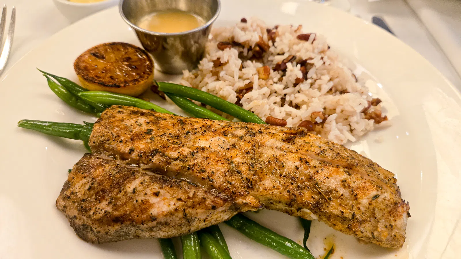 Grilled seasoned fish fillets served with green beans, rice and peas, lemon half, and sauce on a white plate.