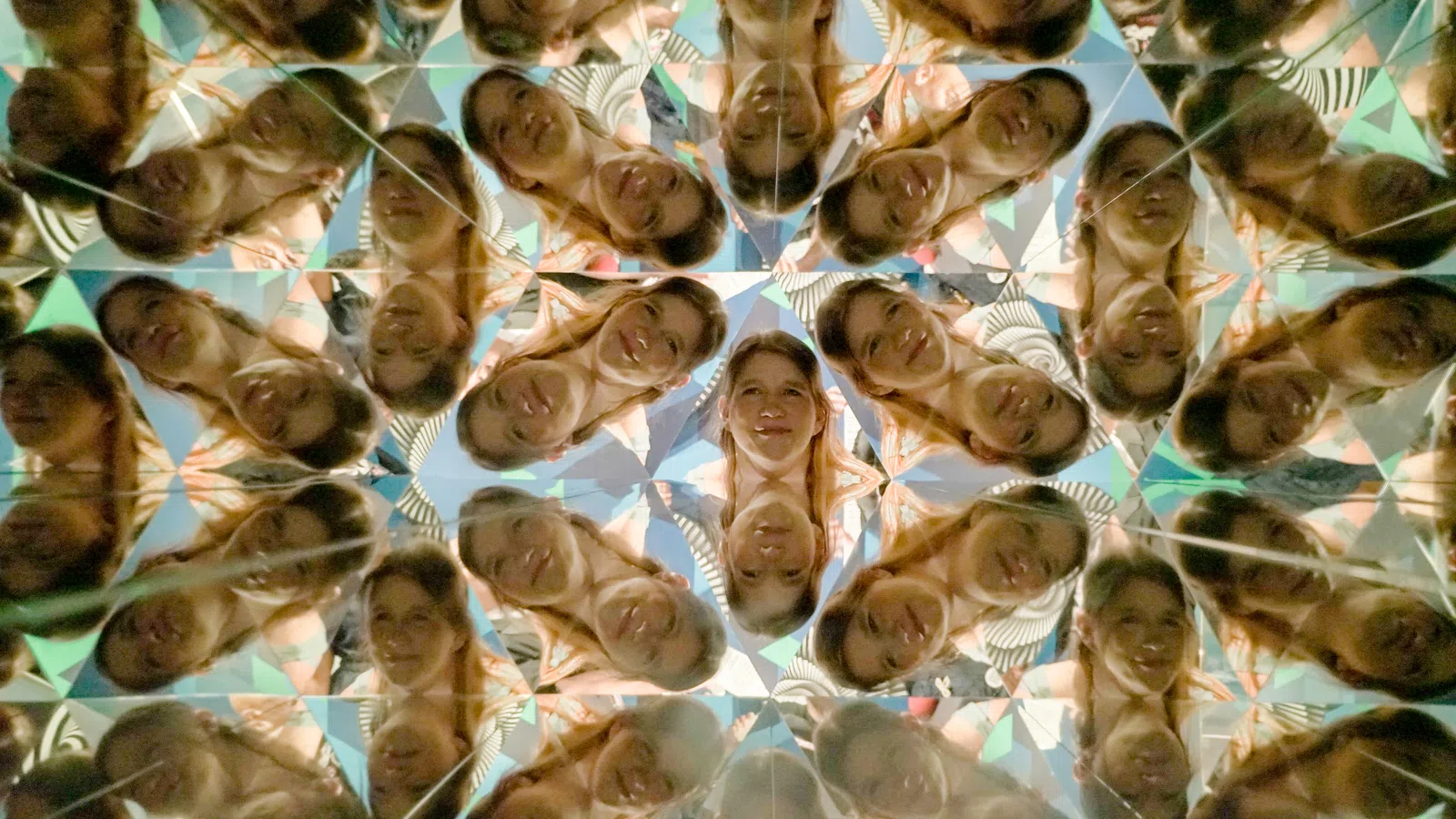 Kaleidoscopic reflections of a smiling woman with brown hair create a complex, colorful pattern of repeating faces.
