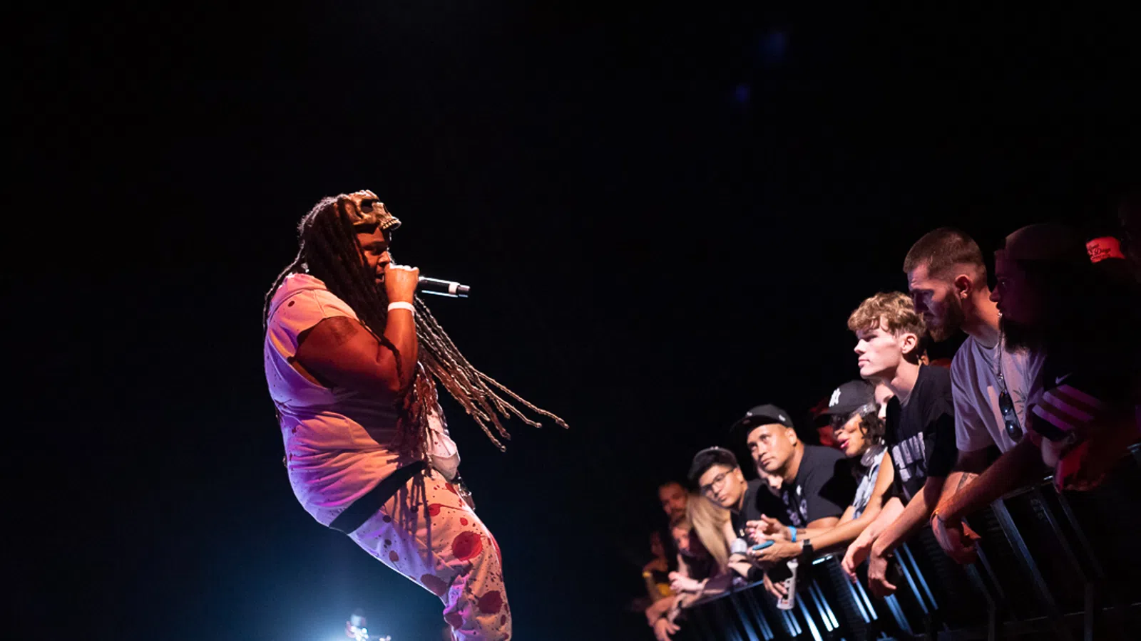 Rapper with long dreadlocks performing on stage with microphone, engaging an attentive crowd in front row