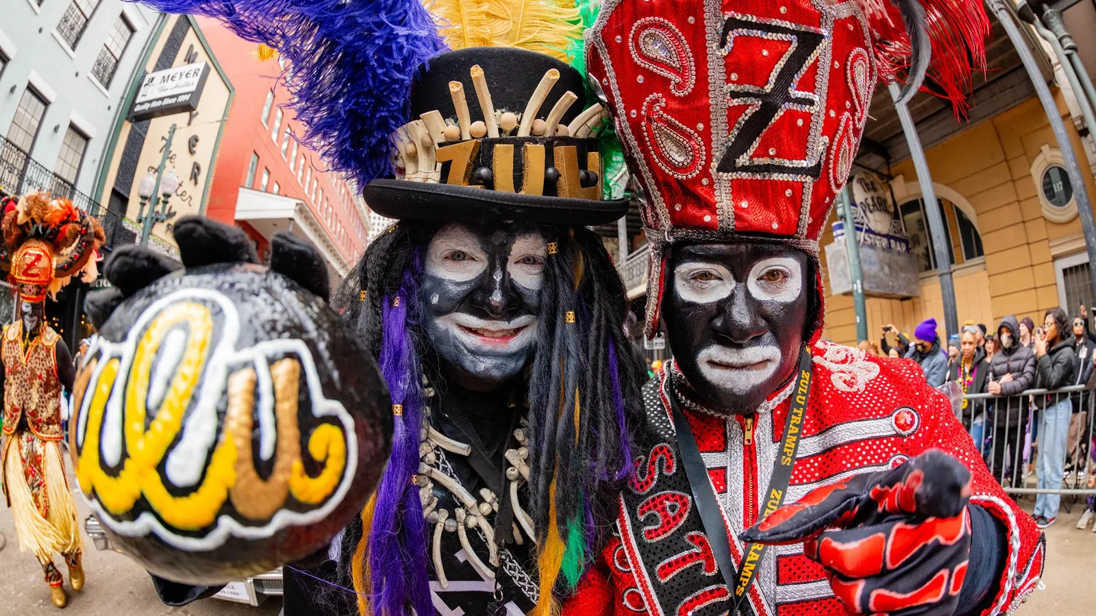 Two Mardi Gras Indians in vibrant, beaded costumes with feathered headdresses at a street parade.