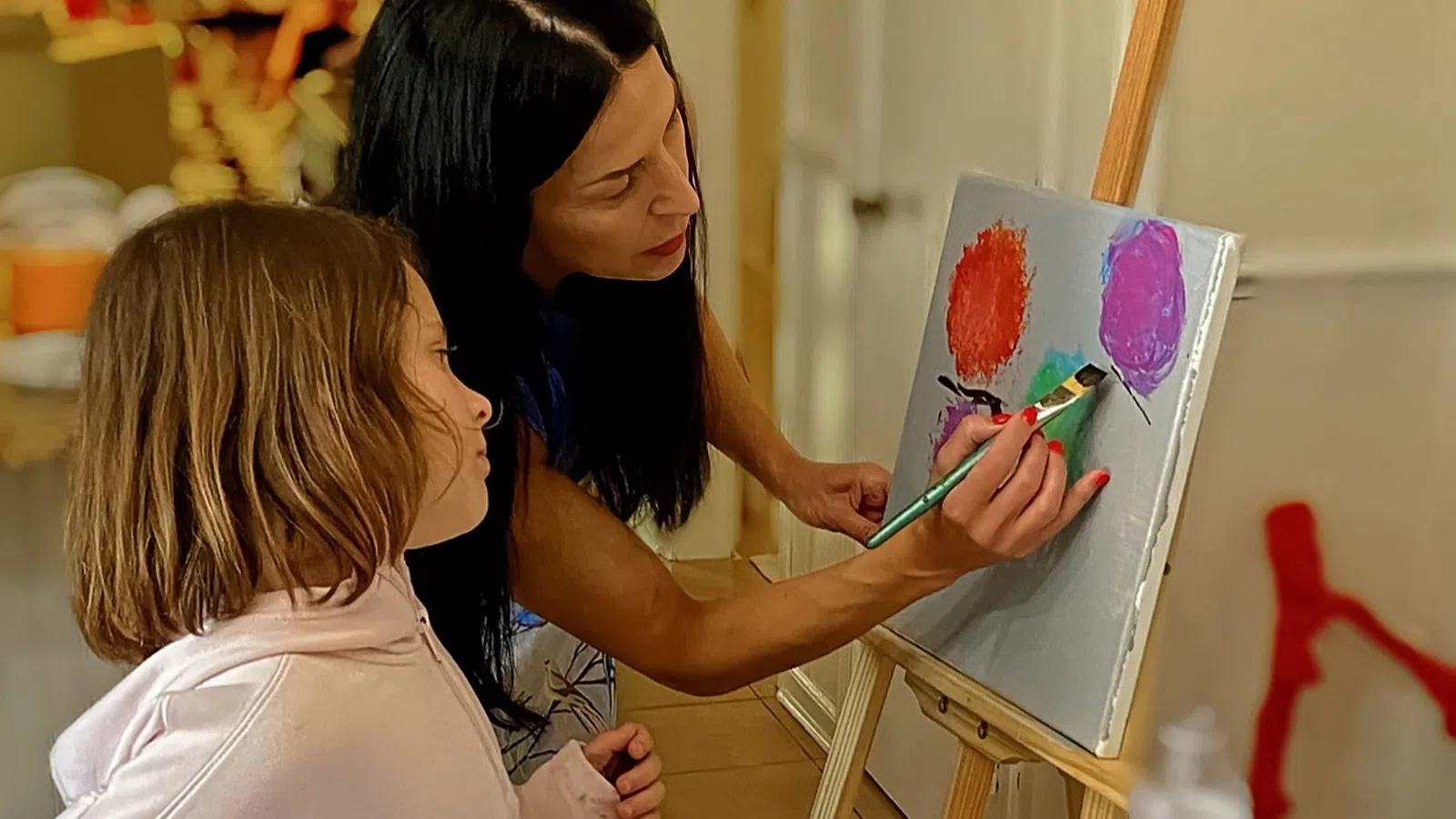 Woman teaching young girl to paint colorful balloons on canvas in indoor art class setting