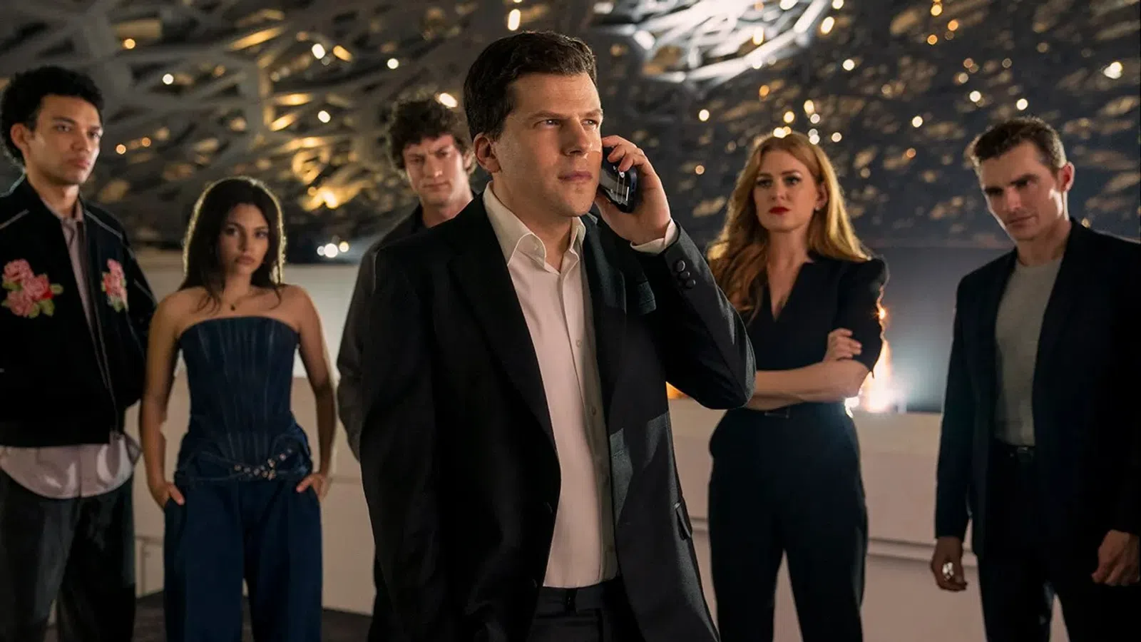 Group of six serious young adults in stylish outfits with one man in the center on a phone call under modern lighting.