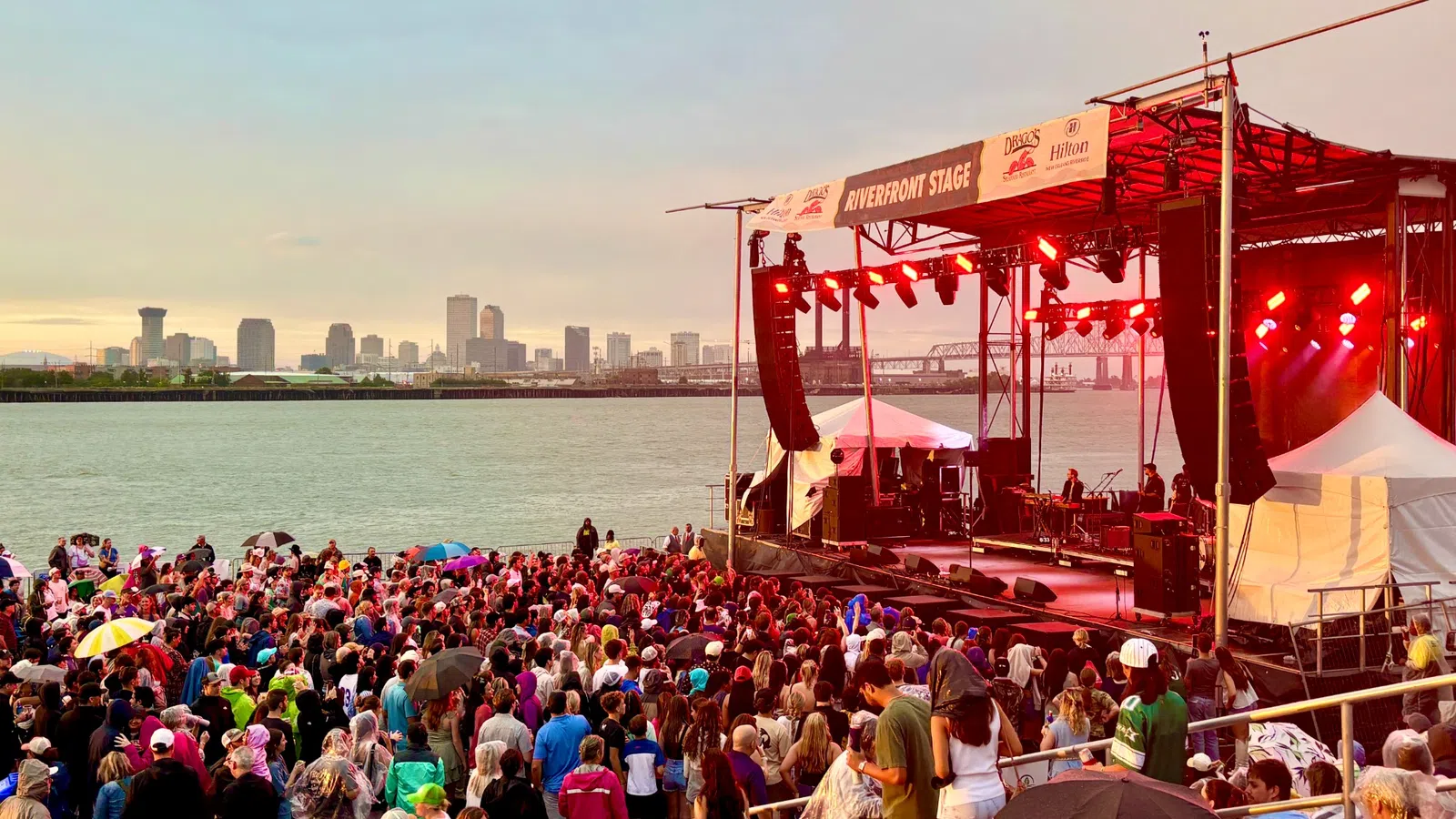 Crowd enjoying live music at the Riverfront Stage with city skyline and river in the background at sunset.