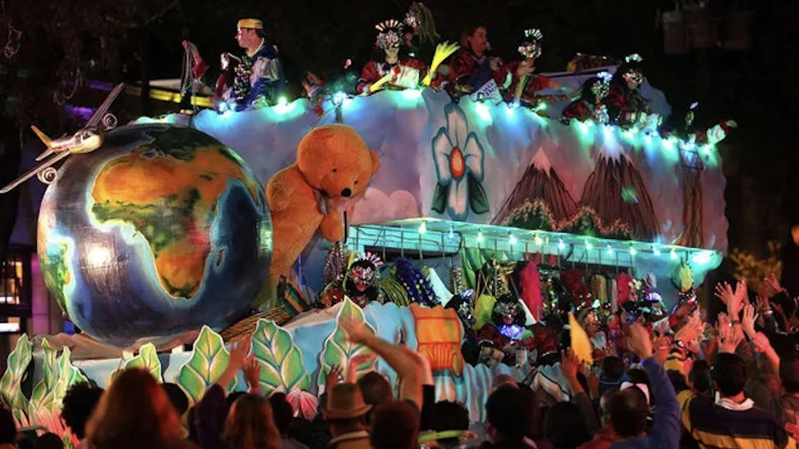 Colorful parade float with large globe, teddy bear, flowers, and people celebrating at night with lights.