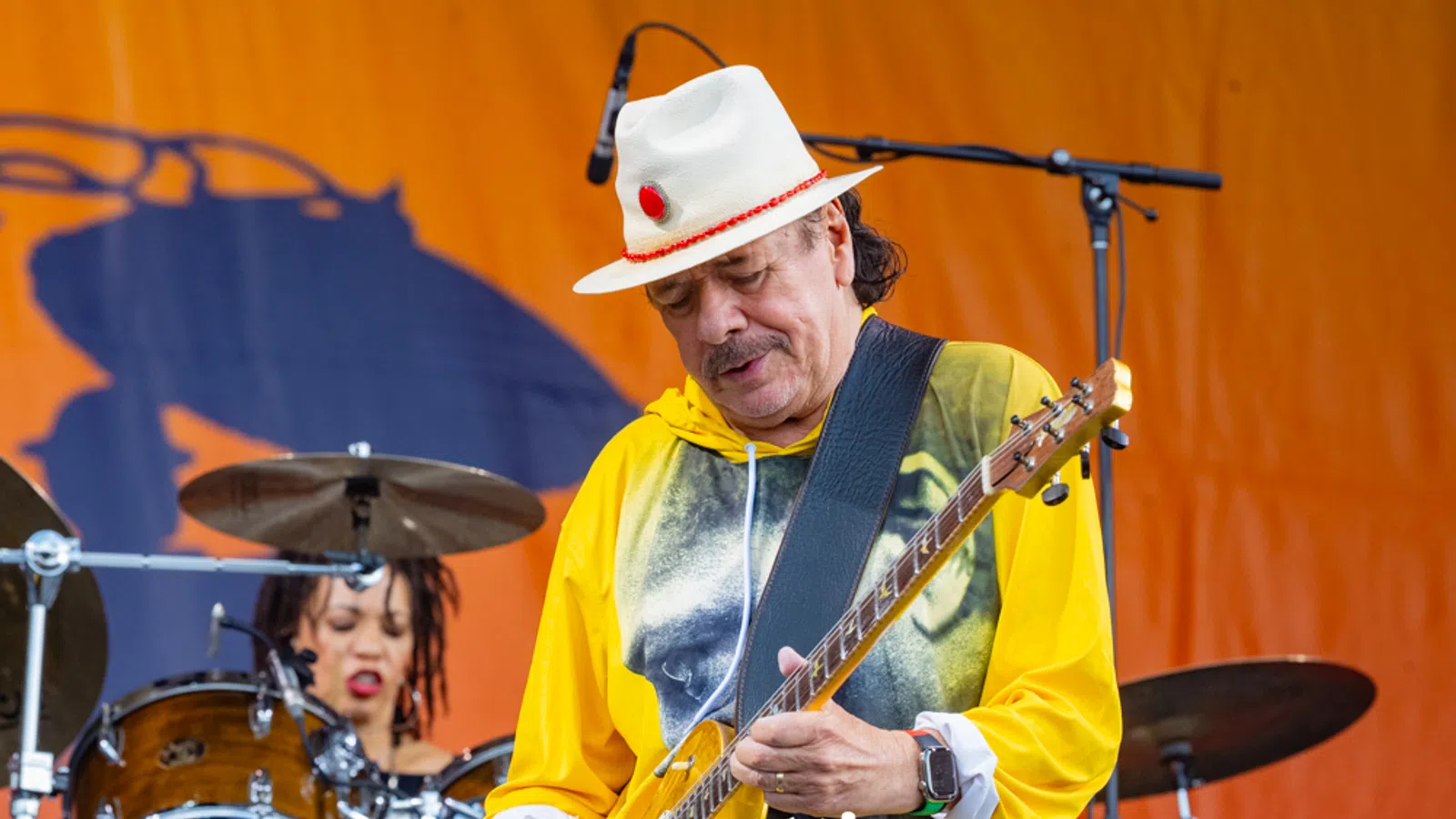 Guitarist in a white hat and yellow hoodie playing on stage with drummer in background against an orange backdrop.