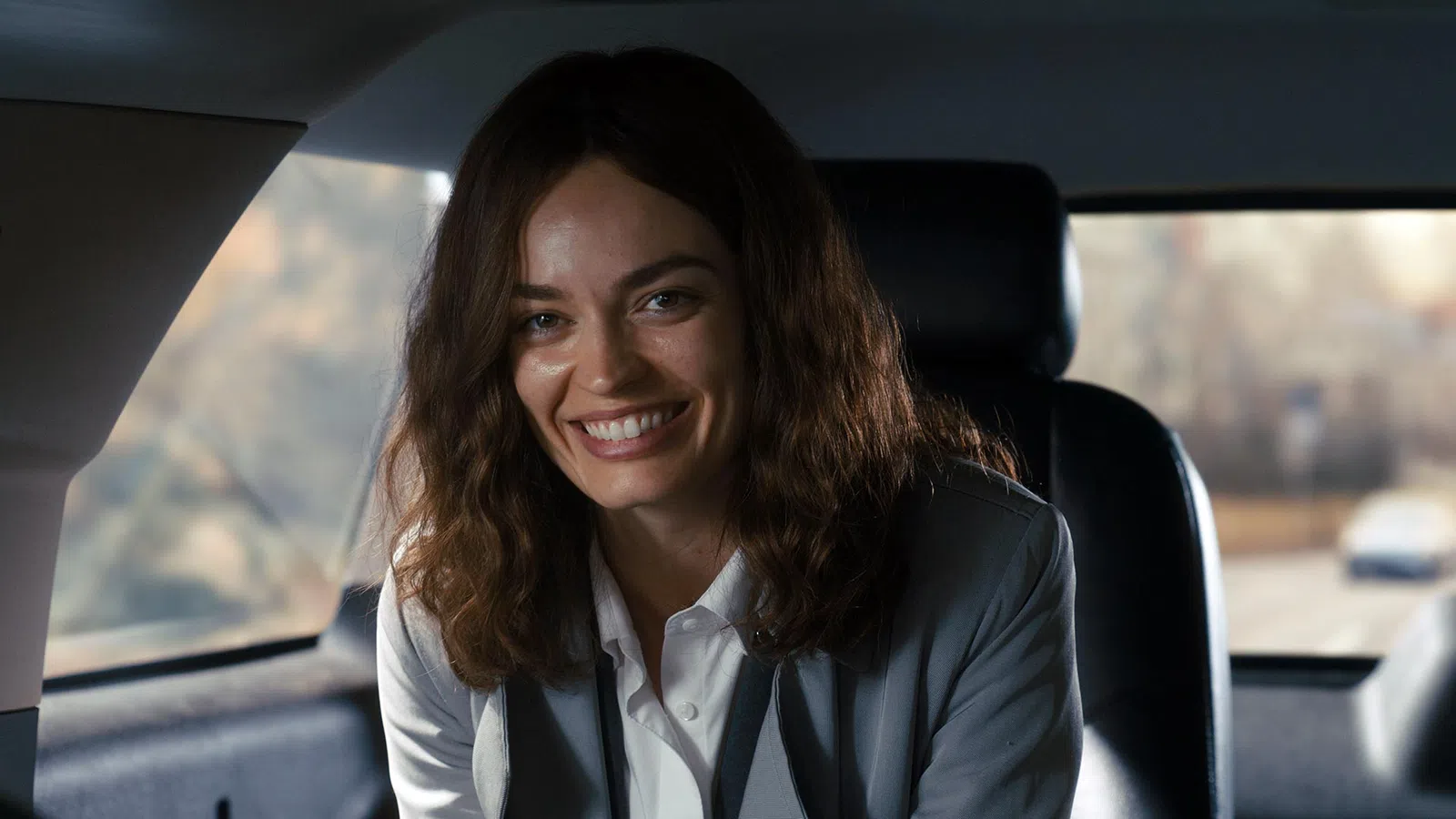 Smiling woman with curly hair wearing a blazer sitting in a car interior with natural light.