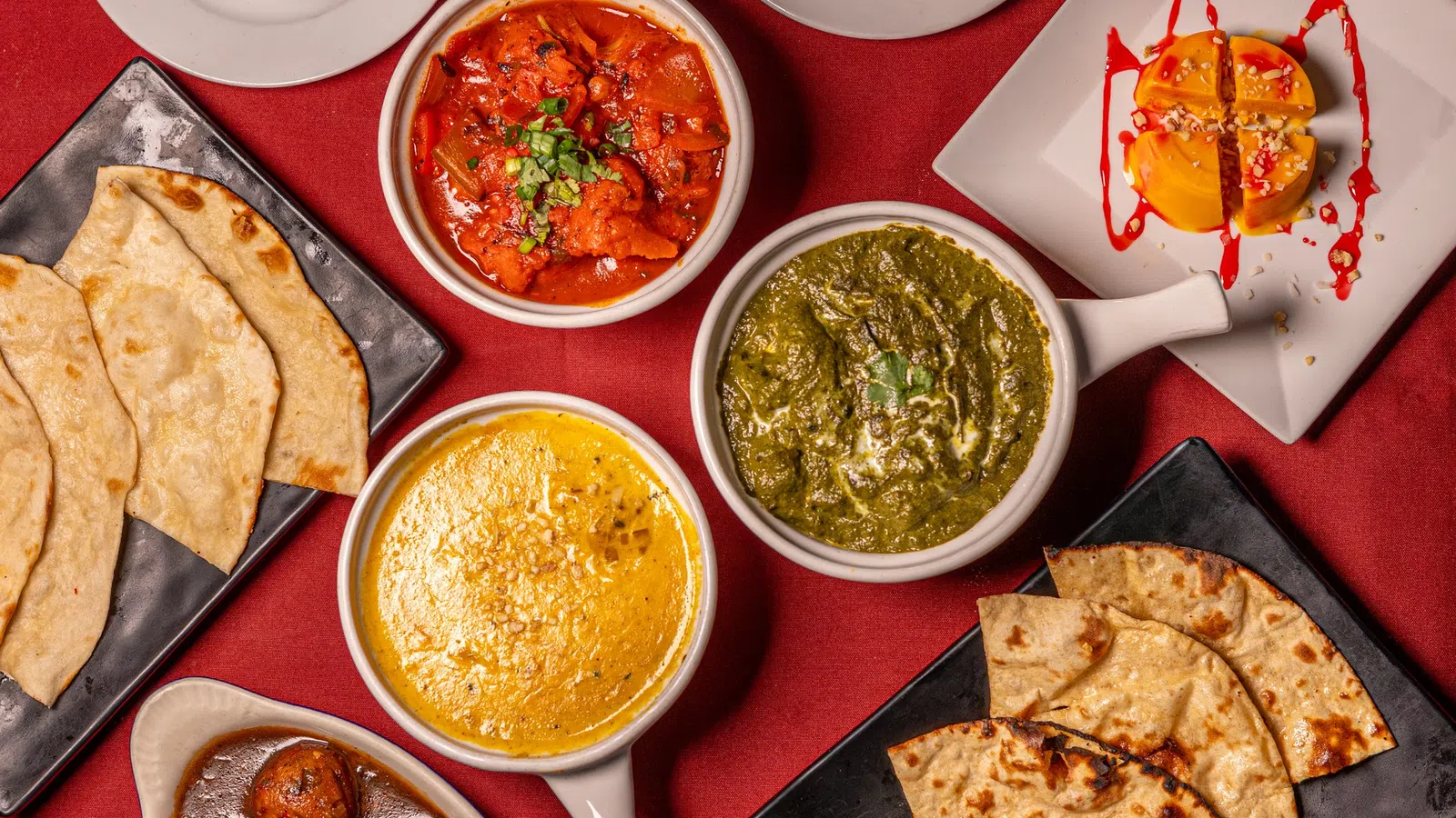Assortment of Indian dishes including naan, paneer curry, spinach curry, gulab jamun, and desserts on red tablecloth.