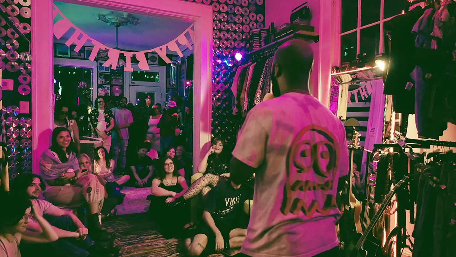 A performer entertains a seated audience in a cozy, decorated room with CDs on walls and hanging clothes.
