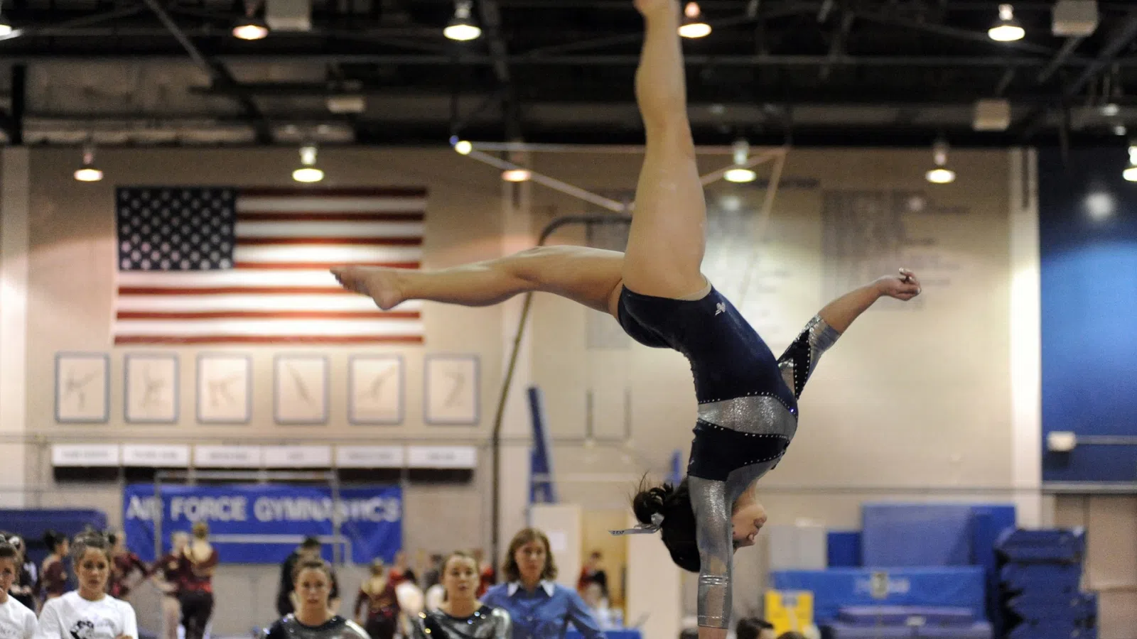 Female gymnast in blue leotard performing handstand routine at a gymnastics competition with US flag backdrop.