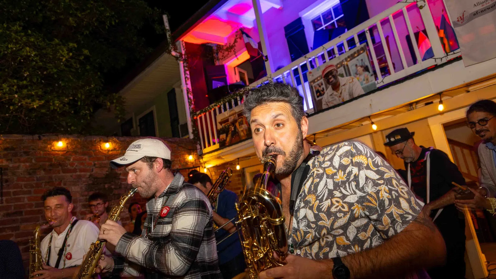Musicians passionately playing saxophones at a lively outdoor night event with colorful lighting and a crowd.