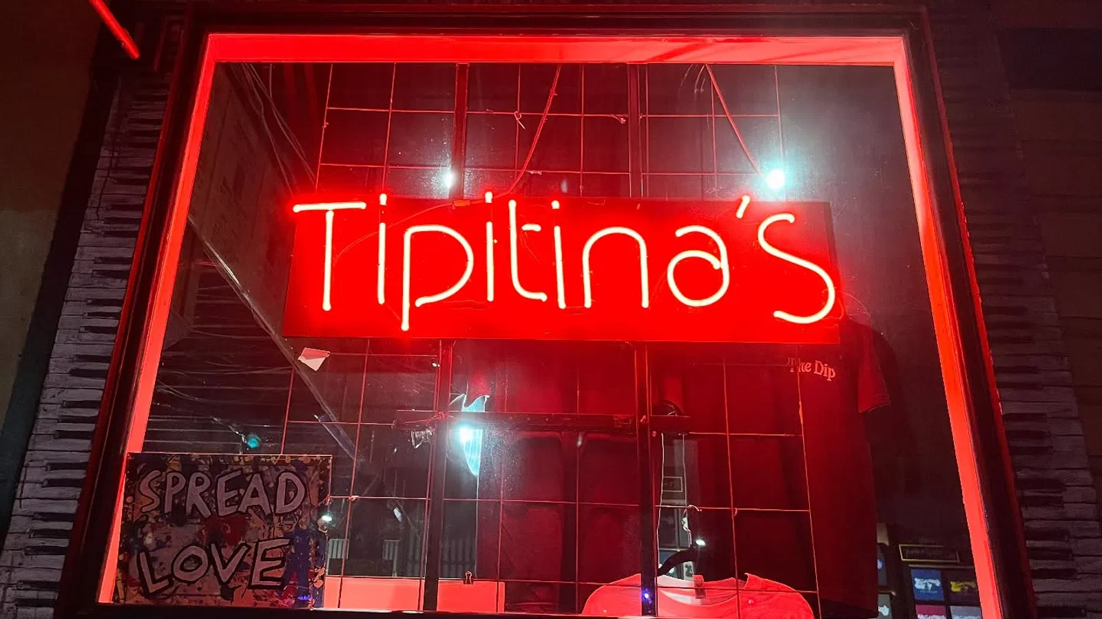 Red neon sign displaying Tipitina's in a window with a Spread Love sign nearby at night.