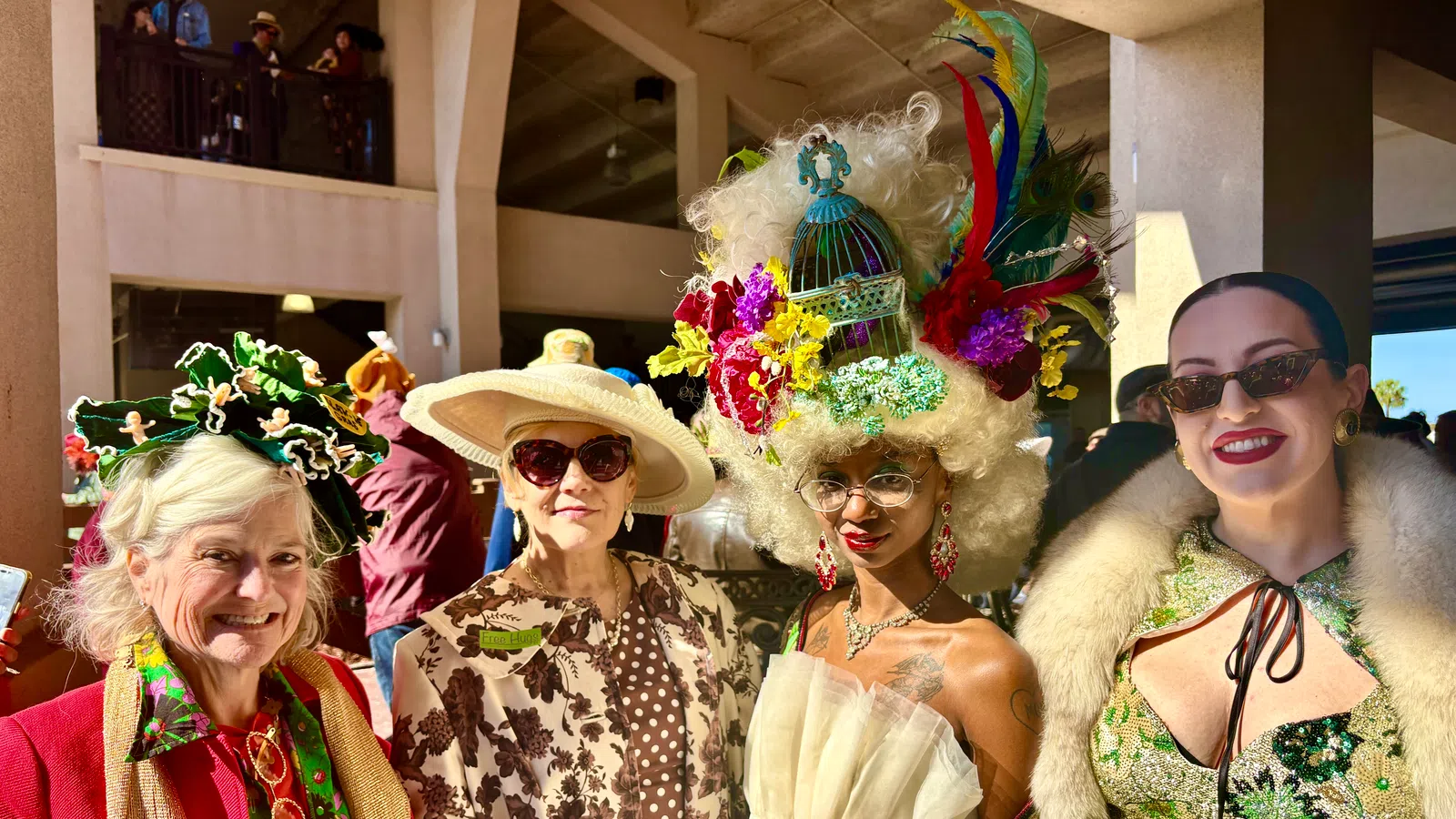 Four women in colorful, extravagant hats and elegant outfits smiling at an indoor event with natural light.