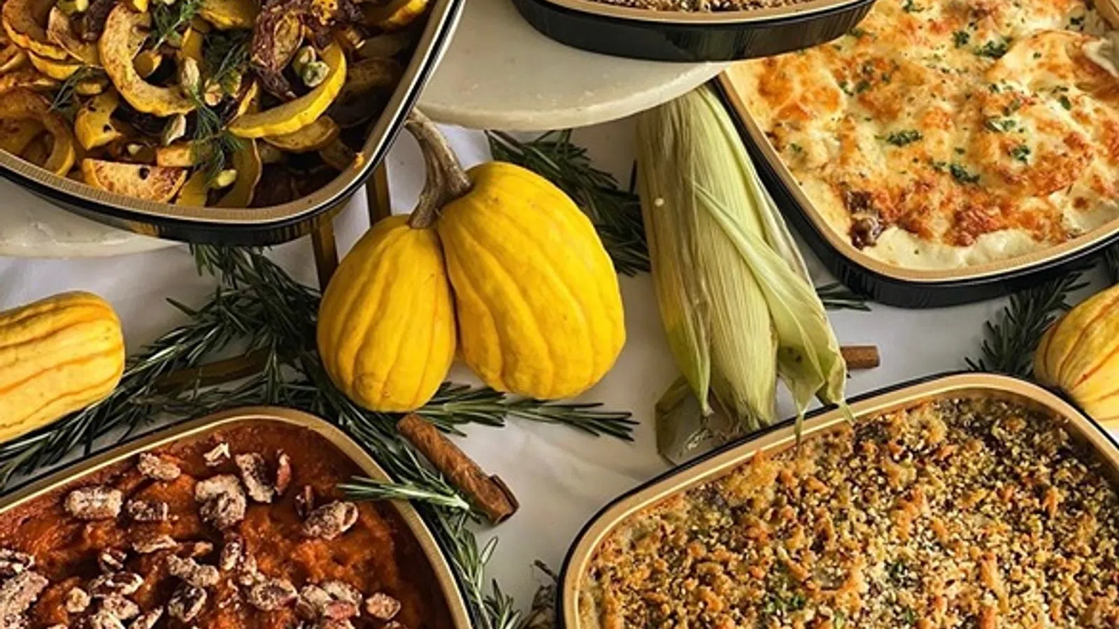 Assorted baked autumn dishes with squash, corn, pecans, and herbs arranged on a white tablecloth