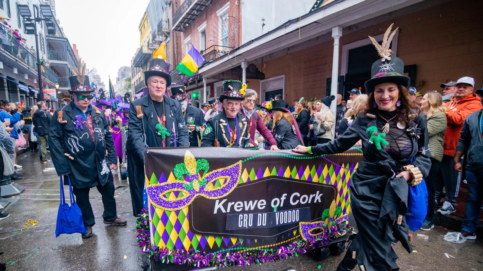 Krewe of Cork members in festive costumes holding a colorful Mardi Gras parade banner on a wet street.