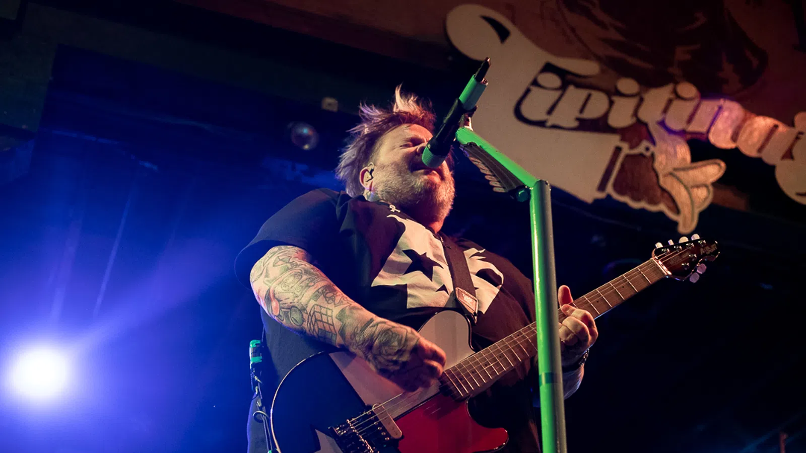 Tattooed musician passionately playing electric guitar and singing into microphone on stage with vibrant lighting.