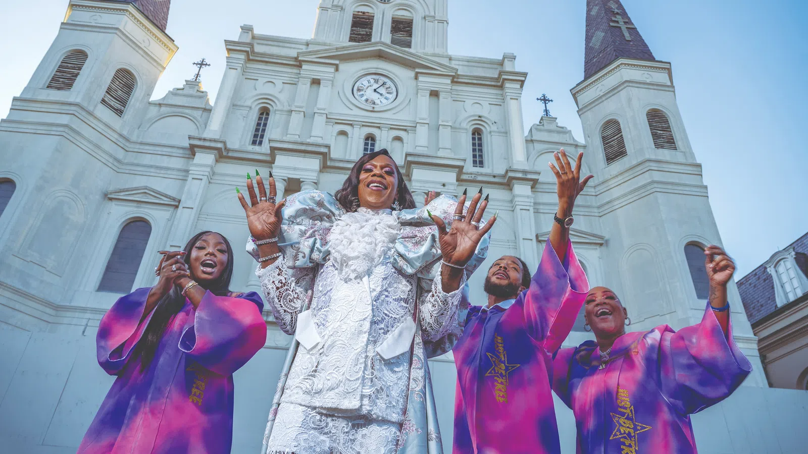 Four joyful singers in vibrant purple and white robes perform in front of a grand historic cathedral with clock tower.
