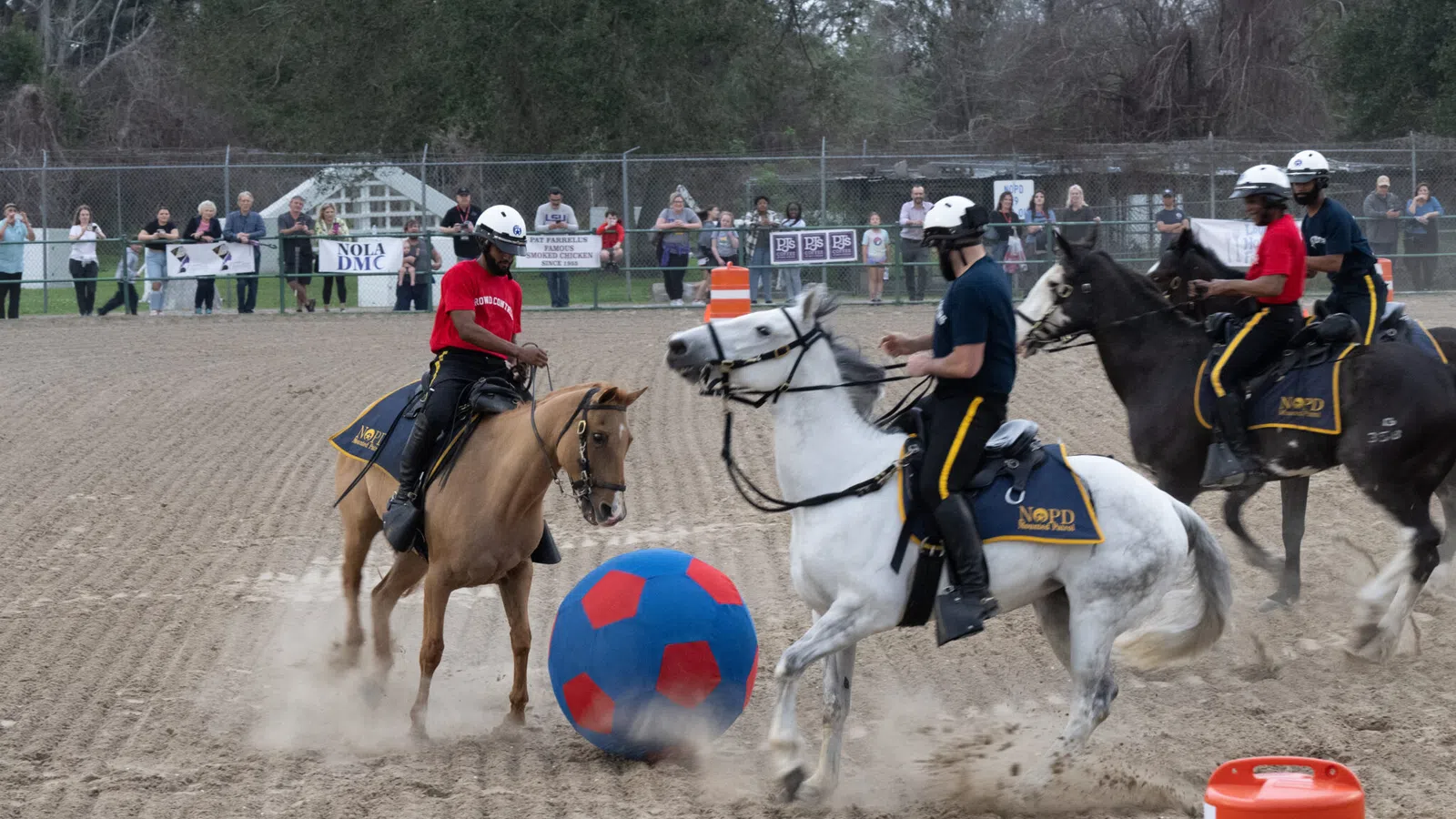 Mounted police officers on horseback play polo with a large blue and red ball on a dirt field with spectators.
