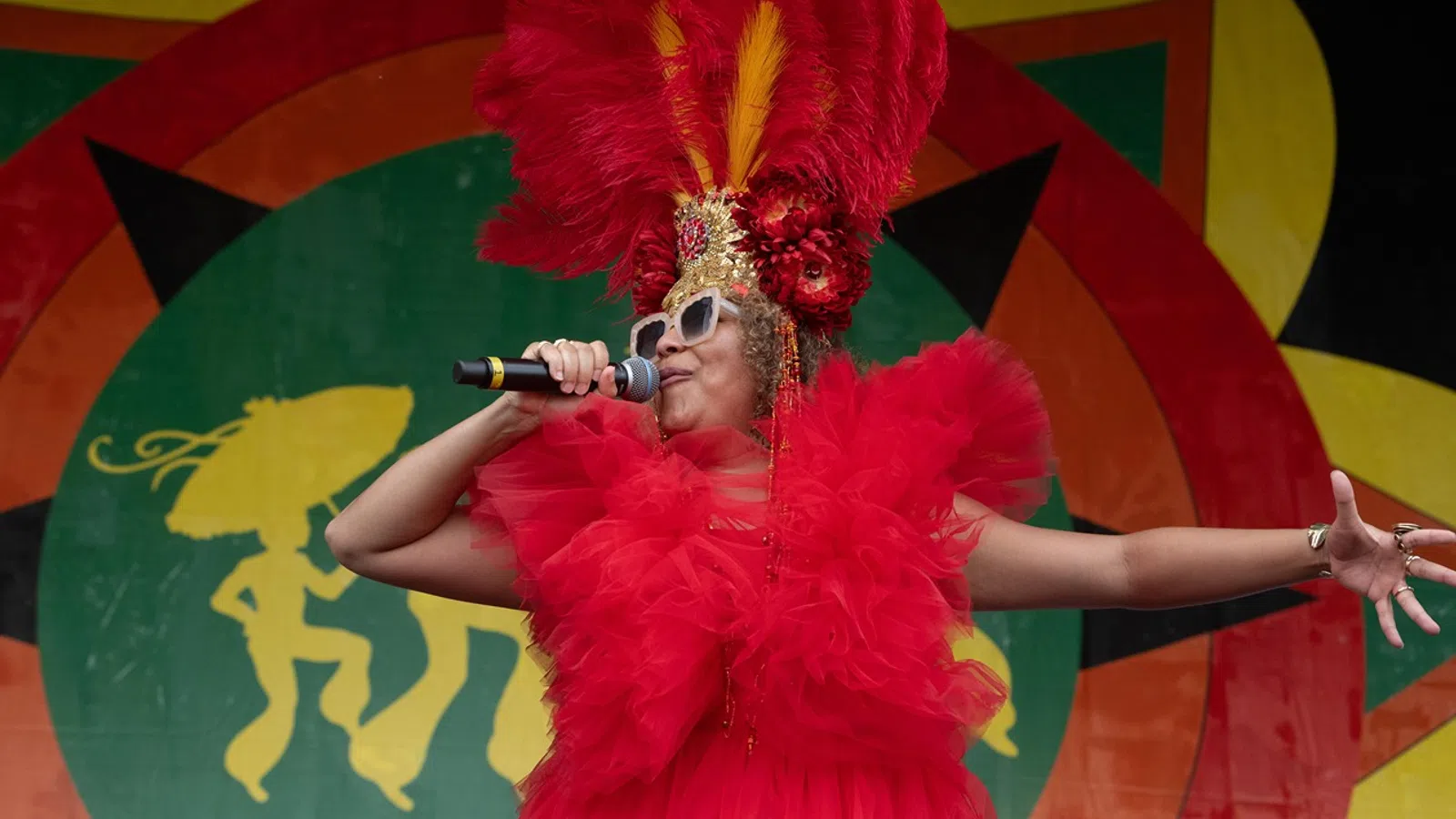 Singer in vibrant red feathered costume and oversized sunglasses performs on stage with colorful backdrop.