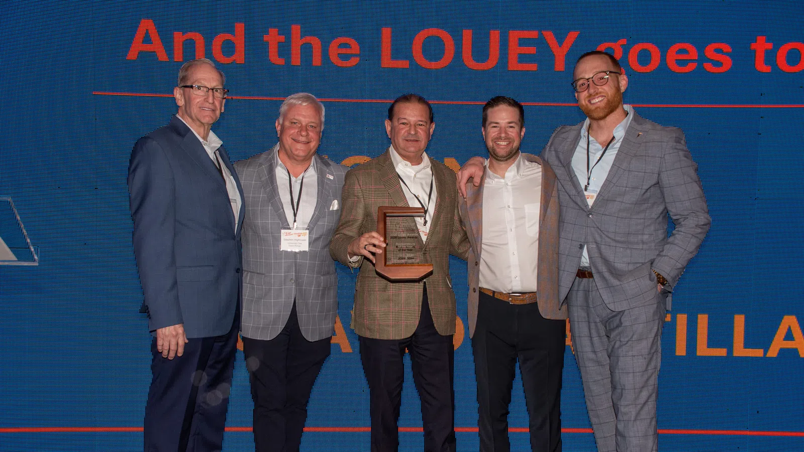 Five men in business attire pose on stage with an award plaque at a corporate event with presentation screen behind them