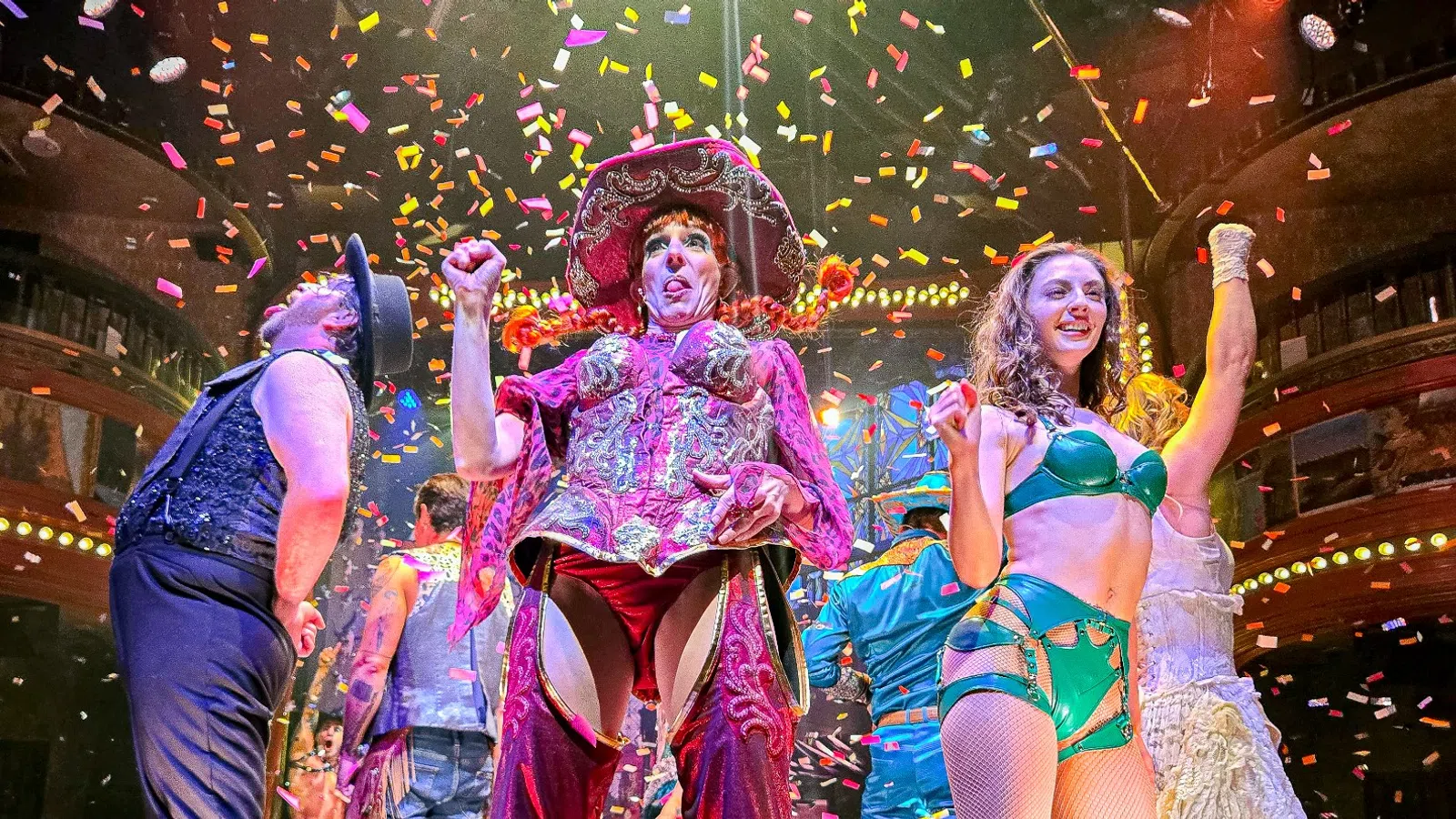 Performers in colorful costumes celebrate on stage with confetti falling and an applauding audience around them.