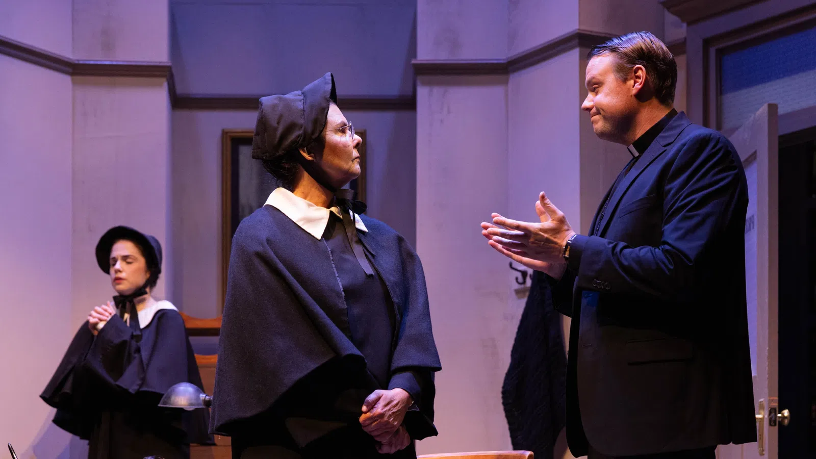 Three actors in period costumes engage in a dramatic scene on stage with serious expressions and historical attire.