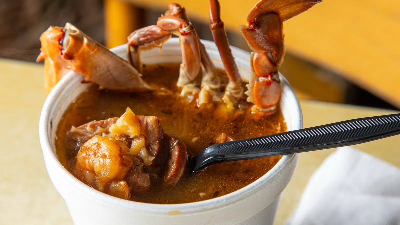 Close-up of a seafood stew with crab legs and shrimp served in a white foam cup with a black spoon.