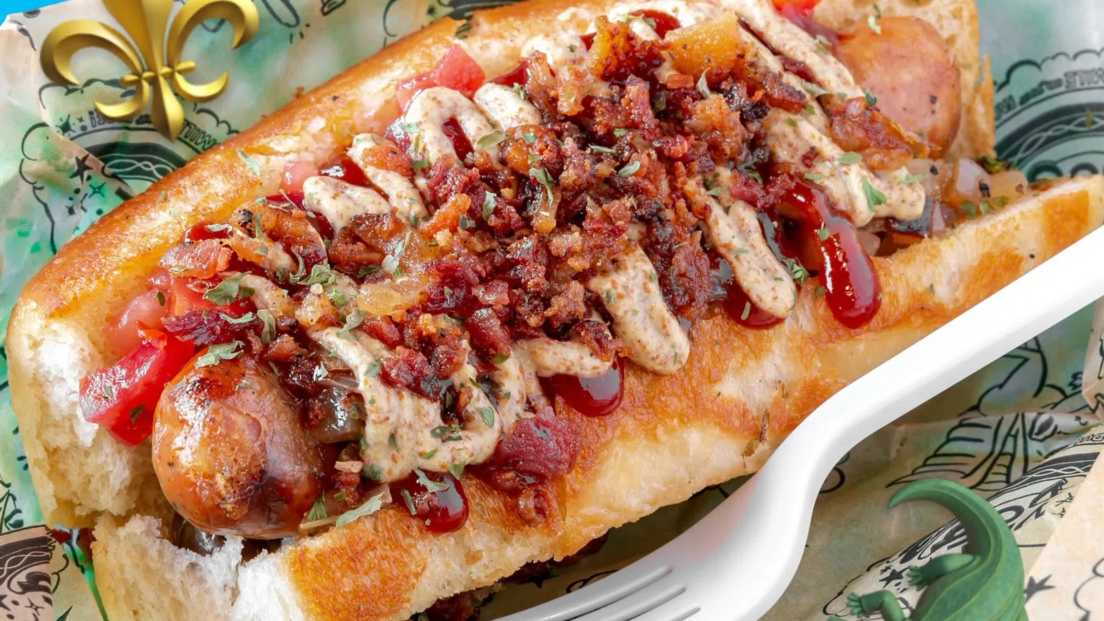 Loaded hot dog topped with crispy bacon bits, sauces, and diced tomatoes in a soft bun on patterned paper.