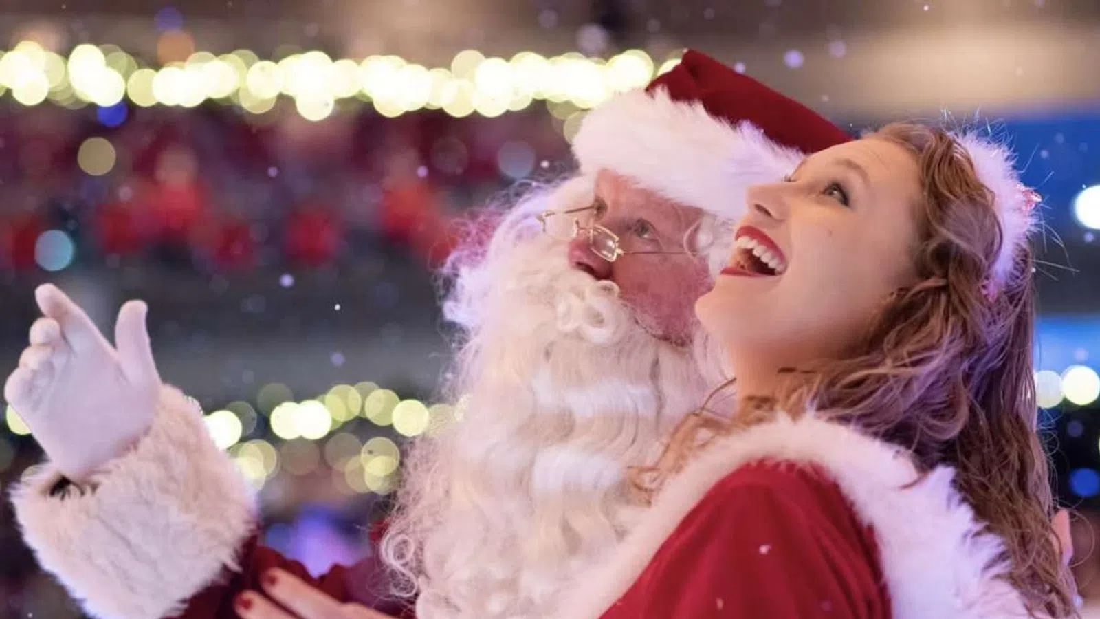Santa Claus and a woman dressed in red joyfully looking up with holiday lights and snowflakes around them.